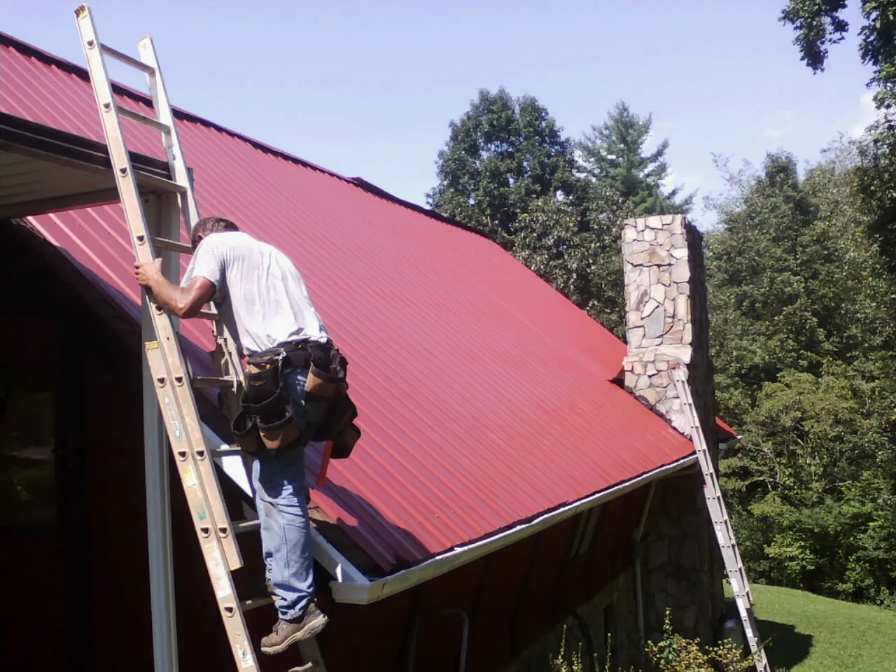 Roofer on a ladder beside a red metal roof and stone chimney — Soffit & Fascia Repair by Roofing Brothers in Camdenton